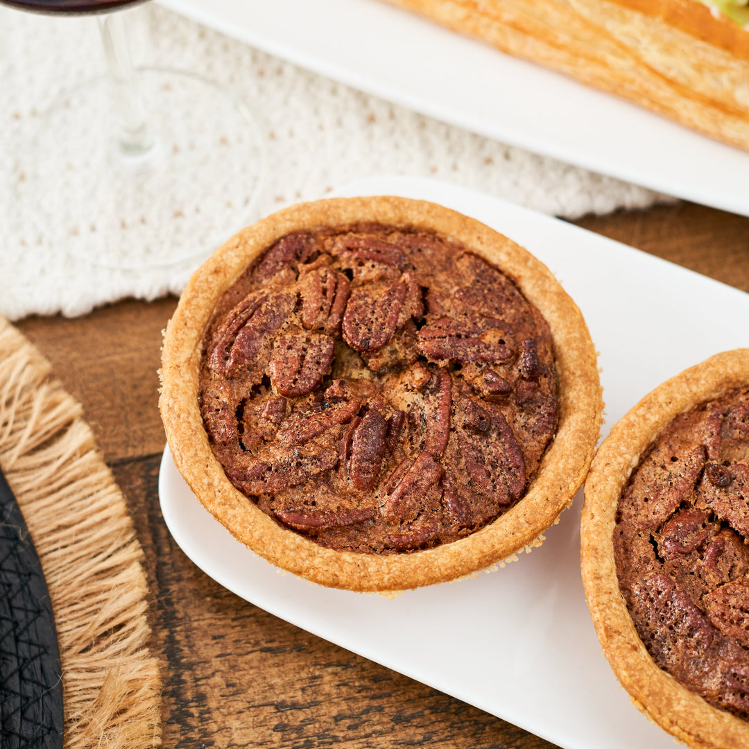 Two pecan tarts on a white plate with a glass of red wine and a sandwich in the background.