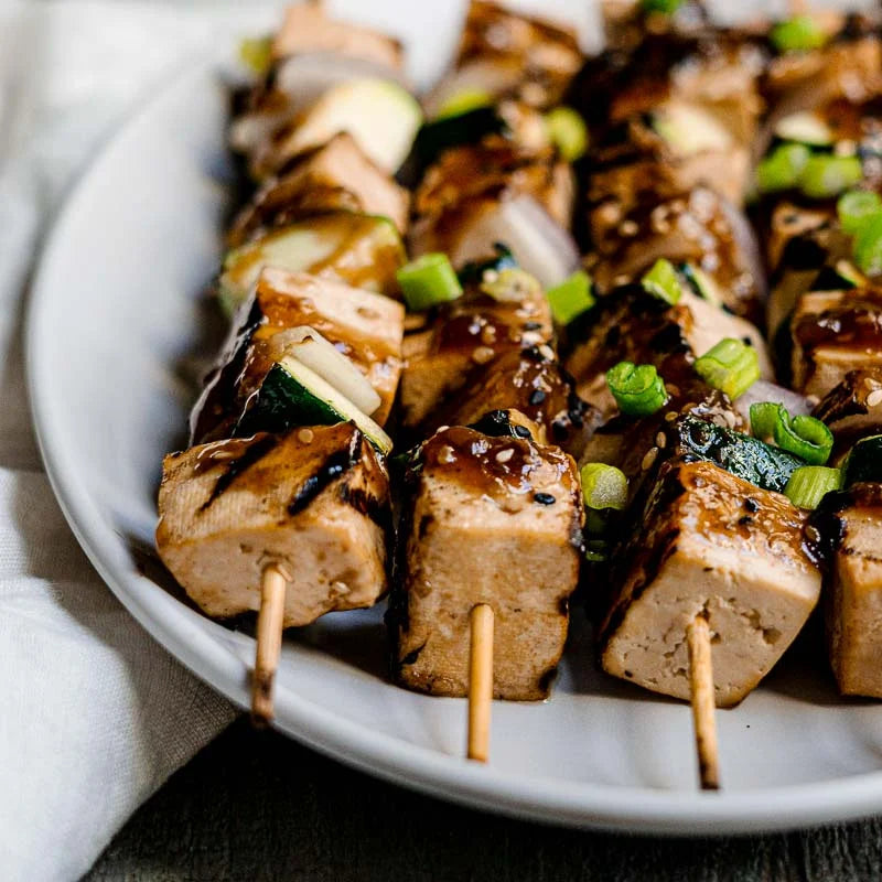 Skewers of tofu and vegetables with a sauce on a white plate.