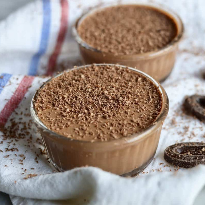 Two small glass cups filled with chocolate pudding on a white cloth with cookies around.