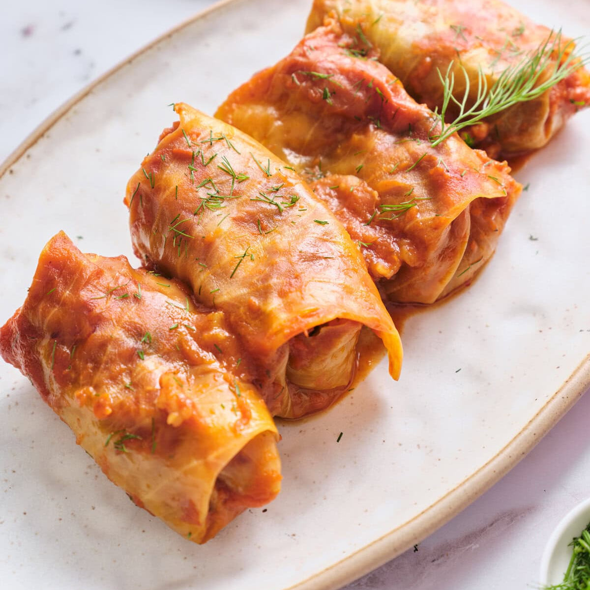 Cabbage rolls on a white plate with a small bowl of sauce in the background