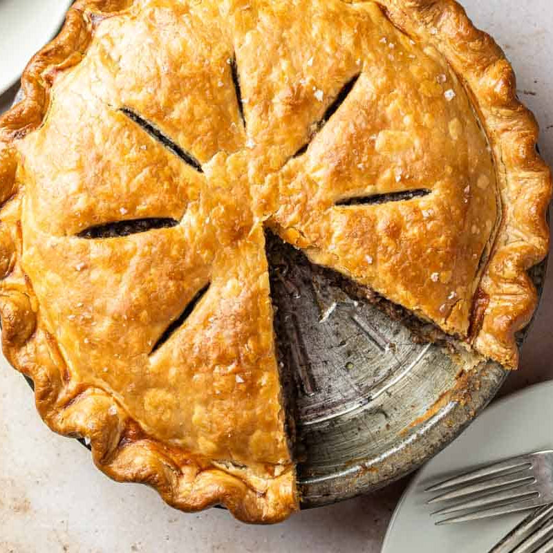 Baked tourtiere with a slice removed on a plate with forks and a glass of tea.